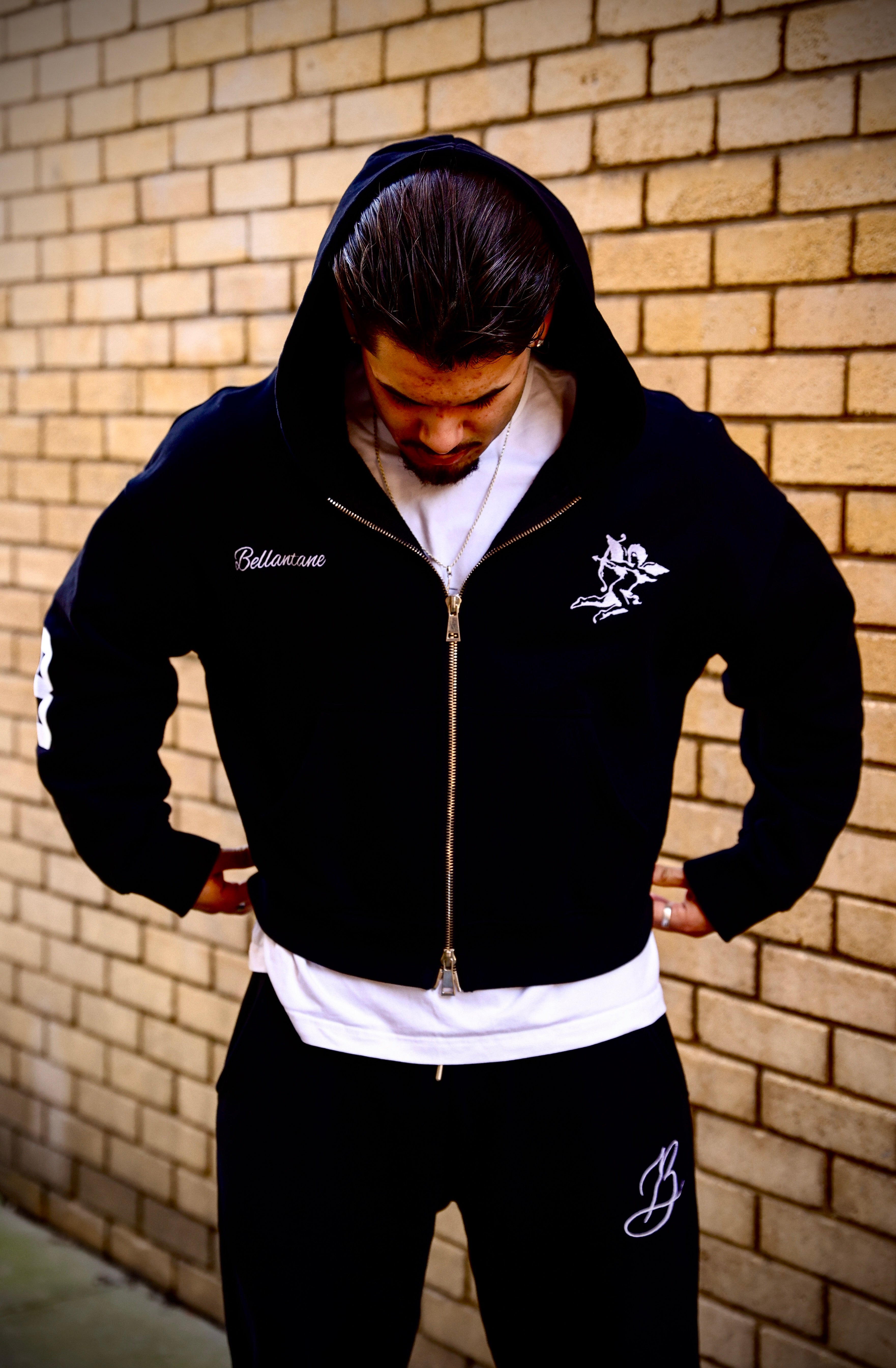 Person wearing a black hoodie and pants with branding against a brick wall.