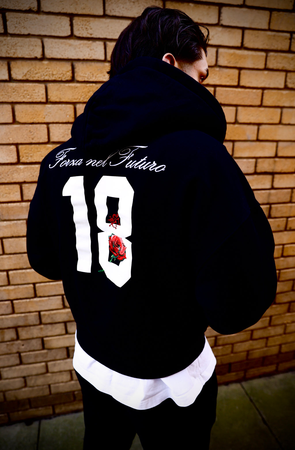 Person wearing a black hoodie and pants with branding against a brick wall with their back facing the camera